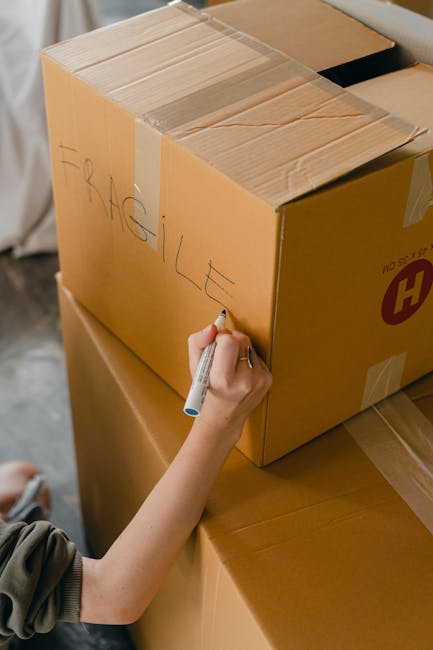 A person in casual clothing, with their face not visible, is lifting a medium-sized cardboard moving box in a well-lit room with hardwood flooring. The individual is using both hands to grasp the box,